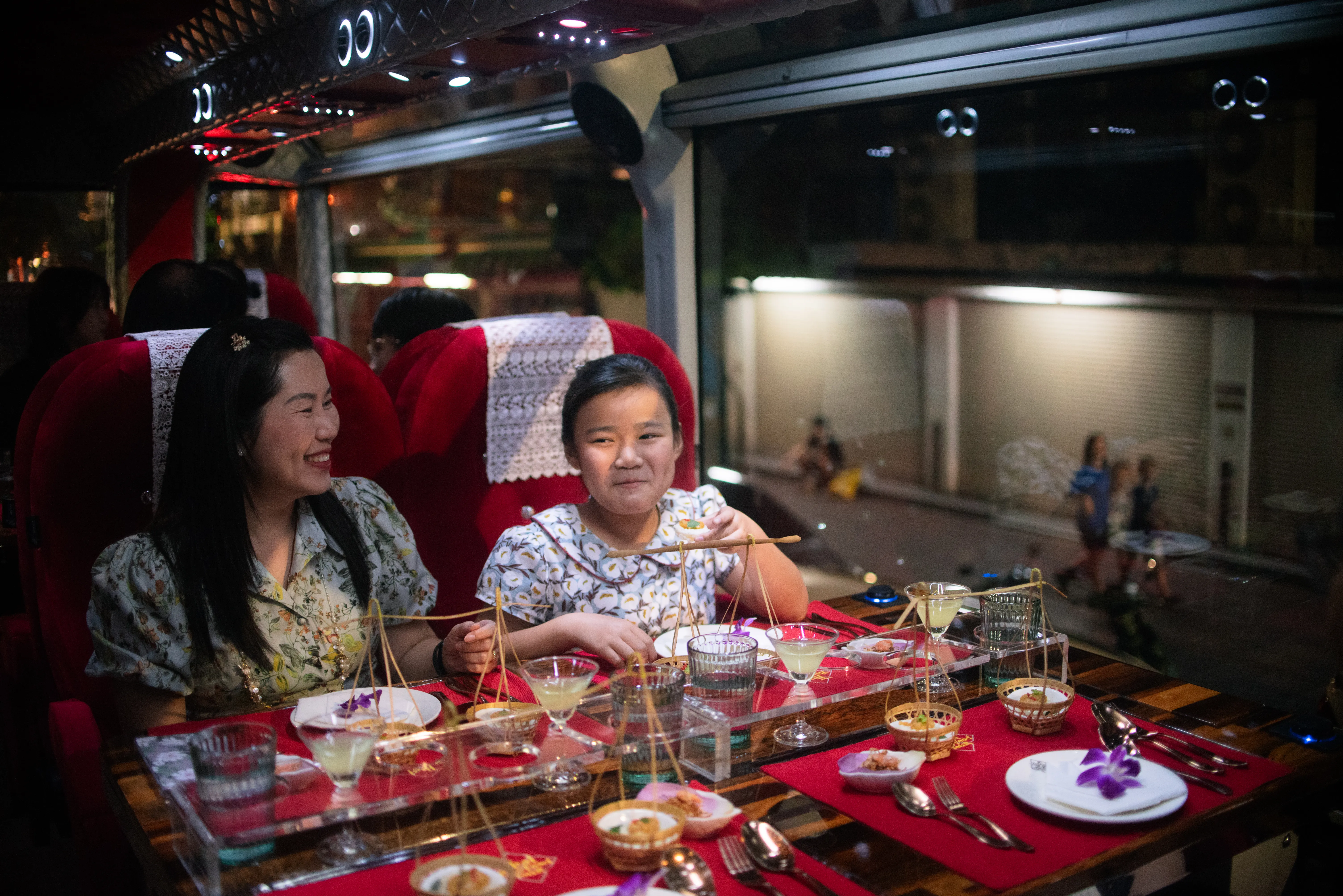 Family with children dining together at a table on the Thai Bus Food Tour luxury double-decker, with Bangkok's Grand Palace visible through the panoramic window