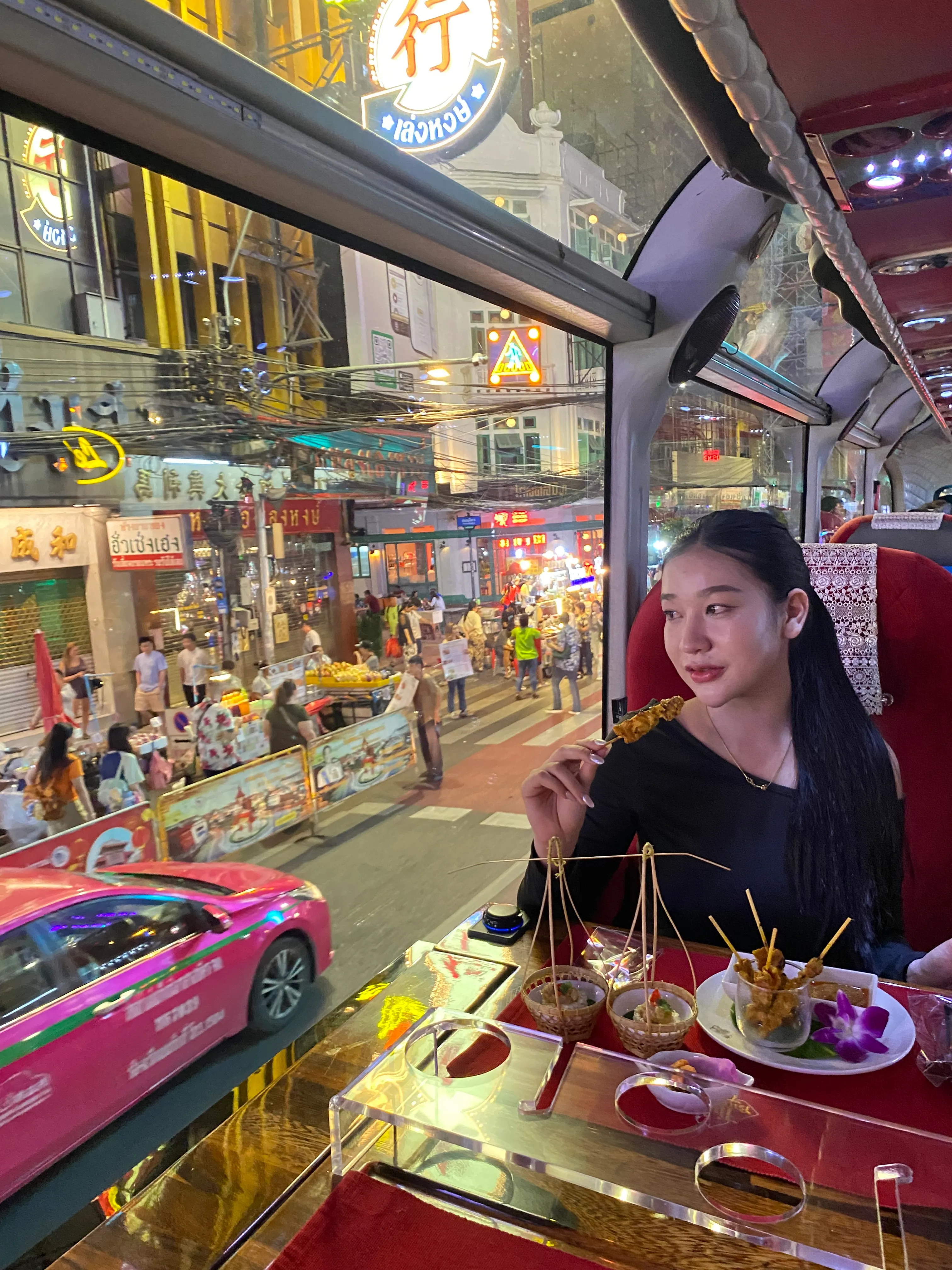 Private group celebrating around tables aboard the Thai Bus Food Tour double-decker at night, illuminated Bangkok landmarks visible through the panoramic windows